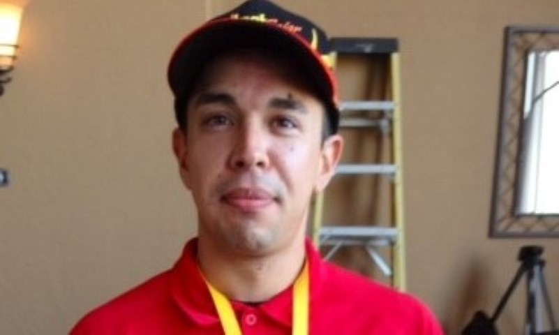 Man in red shirt and cap with yellow lanyard smiles at camera in indoor setting