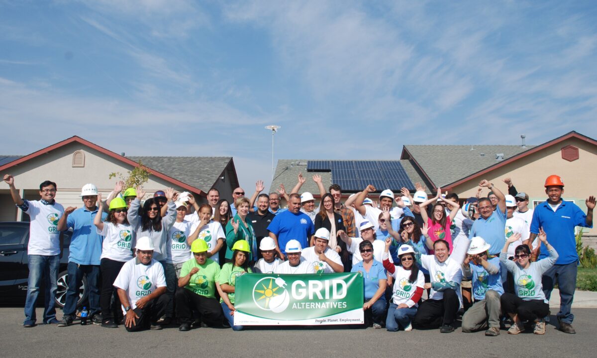 Large group of volunteers wearing hard hats and GRID Alternatives t-shirts holding green banner at solar installation site