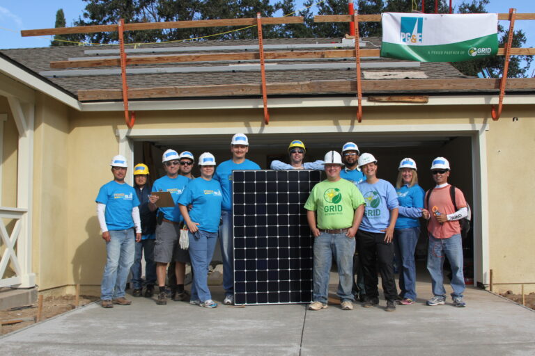 Team of volunteers in blue hard hats holding a large solar panel in front of a house under construction with solar equipment on the roof