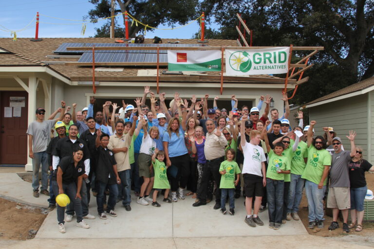 Large group of volunteers with raised hands celebrating in front of house with solar panels and GRID Alternatives banner