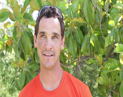 Man in red shirt smiling outdoors surrounded by green foliage