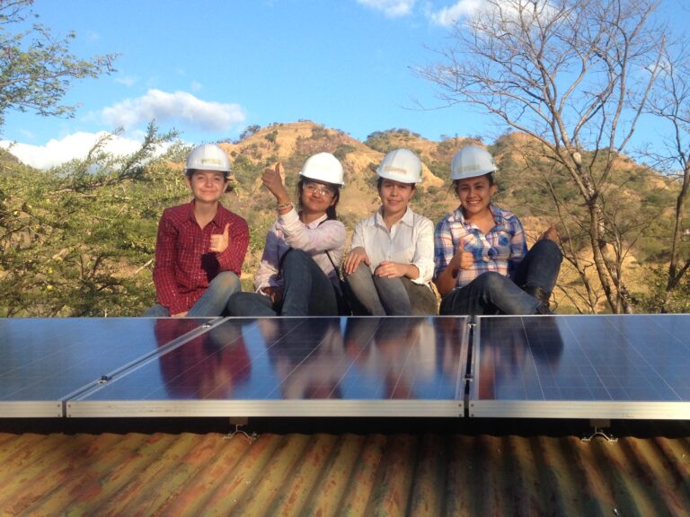 Four women in hard hats sit on solar panels with hills in background, representing women in renewable energy