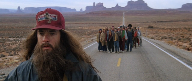 Man with long beard and red cap stands on desert highway with group of people in distance