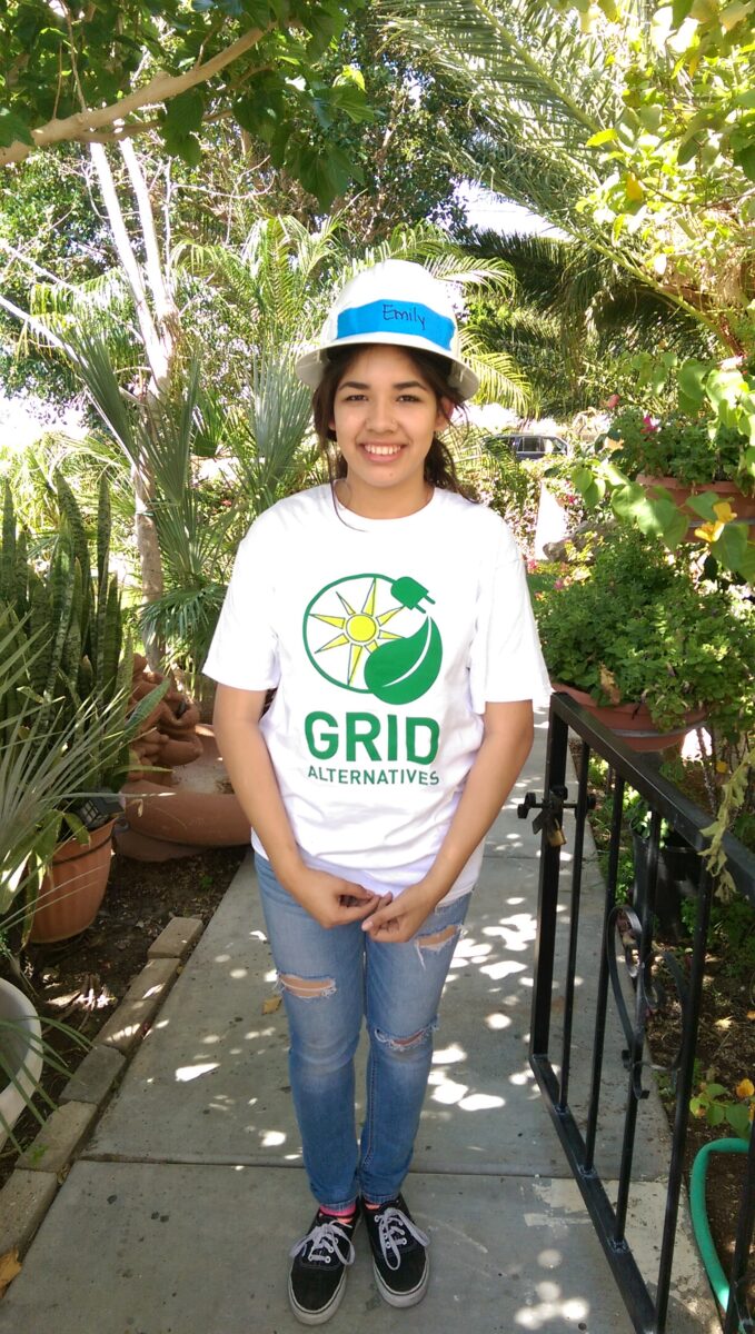 Person wearing GRID Alternatives t-shirt and blue cap smiling in a lush garden setting