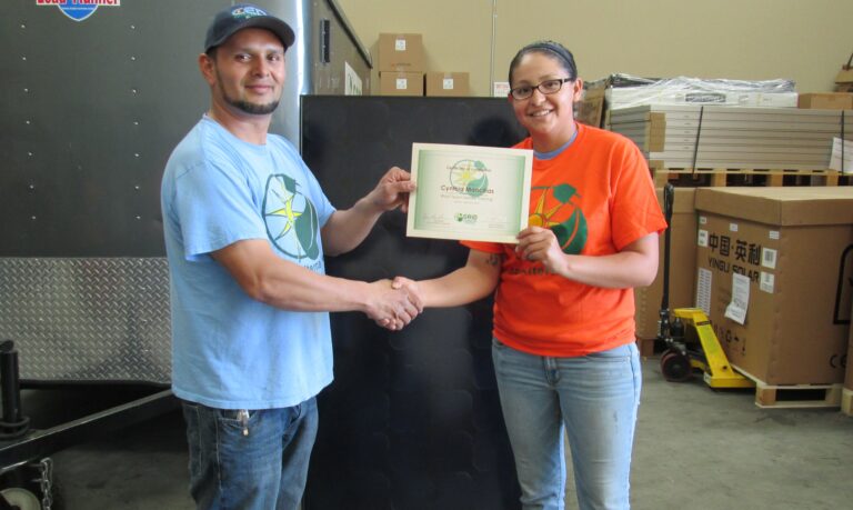 Two people shake hands while holding a framed Cymbal Musicals certificate in a warehouse setting