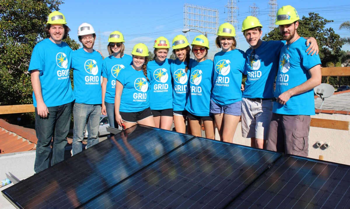 Team wearing blue GRID Alternative shirts and hard hats posing behind solar panels during UNC Solar Spring Break 2015