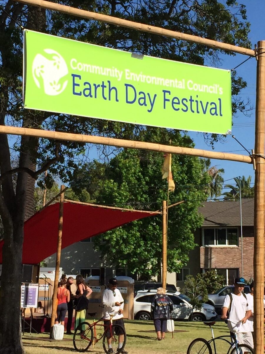Bright green banner reading "Community Environmental Council's Earth Day Festival" at outdoor event with attendees and bicycles