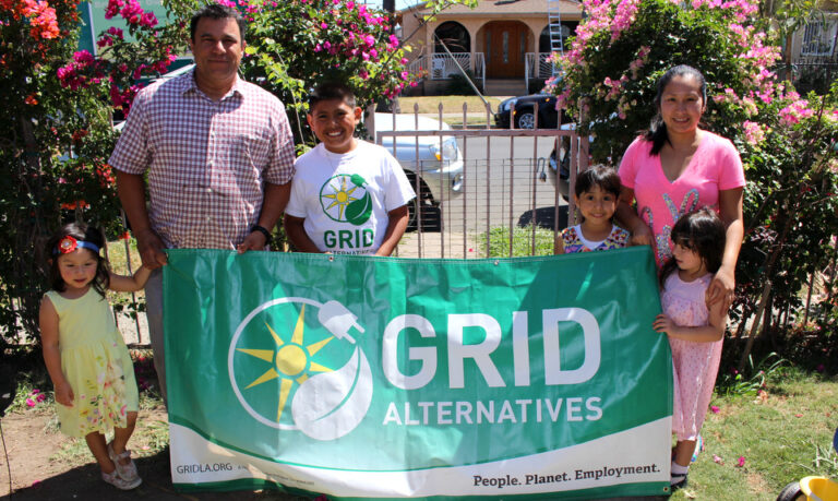 Family holding GRID Alternatives banner with sun logo in front of blooming magenta flowers and house