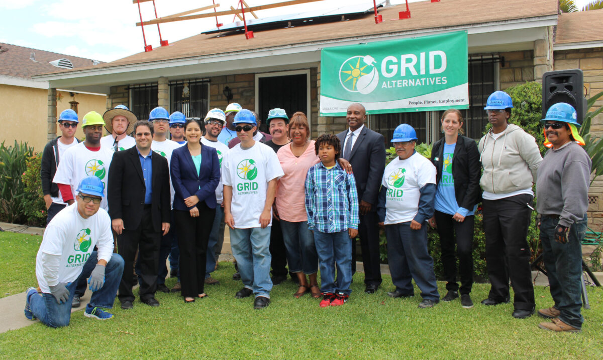 Grid Alternatives team and workers pose in front of a house with solar panels under construction, with green GRID Alternatives banner