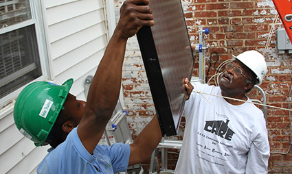 Two workers install solar panel components on a brick house exterior