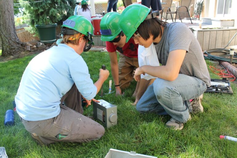 Three young people wearing green hard hats crouch on grass examining electrical equipment during hands-on STEM training