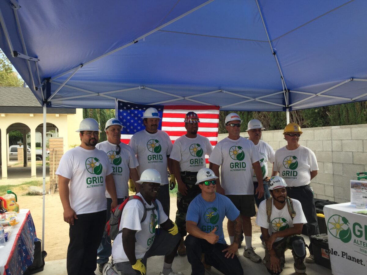 Group of workers in GRID Alternatives shirts and hard hats under blue tent with American flag