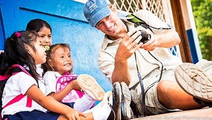 Adult in work clothes showing tool to two smiling children outdoors