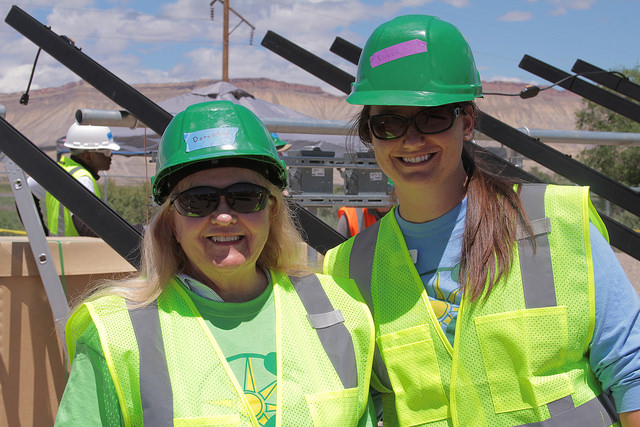 Two women in green hard hats and safety vests smile at a solar panel installation site