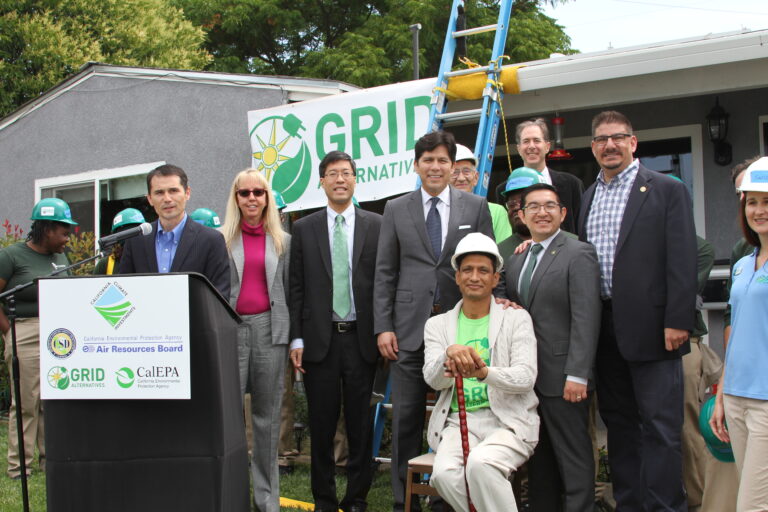 Group of officials and workers at GRID Alternatives solar installation event with podium and blue ladder