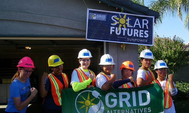 Seven workers in hard hats and safety vests hold a GRID Alternatives banner under a Solar Futures Sunpower sign at a solar installation site