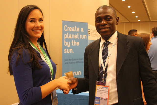 Two professionals shaking hands at a job fair event with "Create a planet run by the sun" signage visible behind them