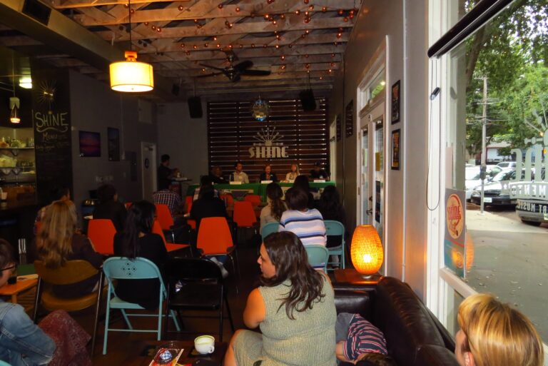 Women seated indoors at a panel event with "SHINE" signage visible at the front table