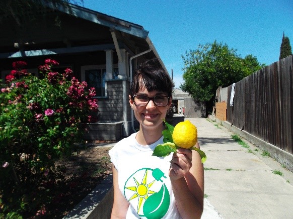 Person holding a lemon in front of a house with pink flowers and clear blue sky