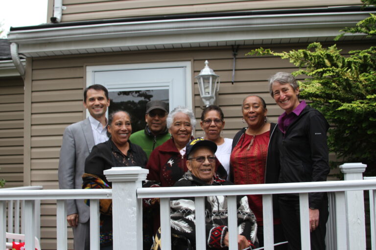 Seven people smiling on a white deck railing in front of a tan house with green ivy