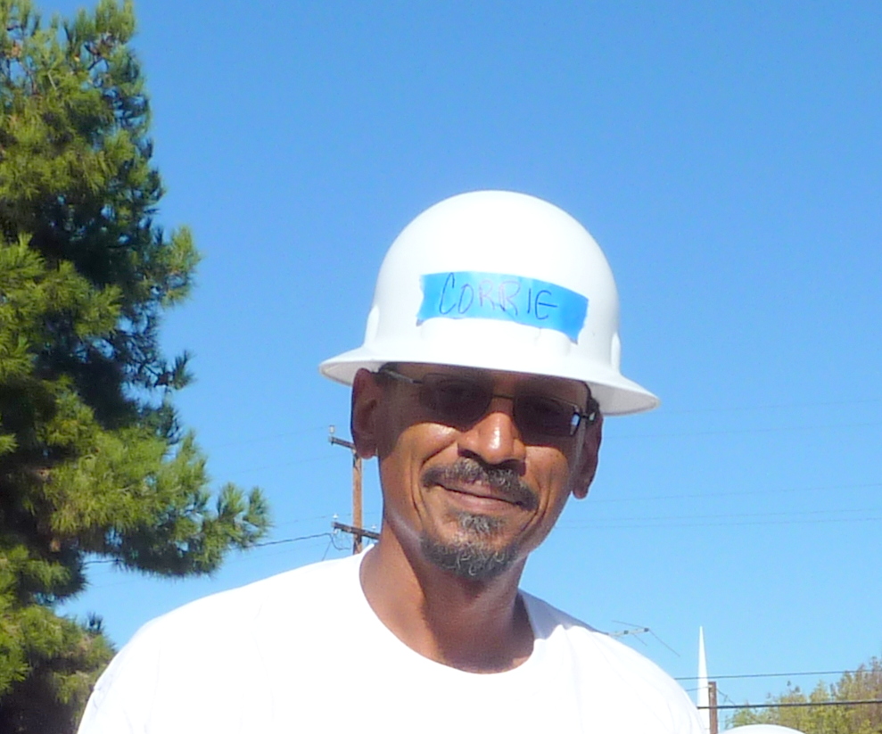 Man wearing white hard hat with "COMING" written on blue tape, sunglasses, and white shirt against clear blue sky