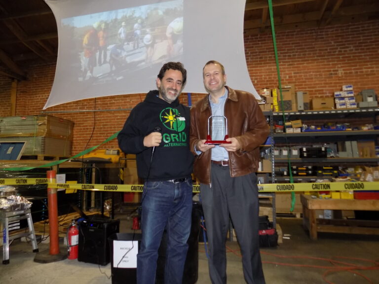 Two men holding an award in an industrial workshop with "GRID Alternatives" banner visible on the wall