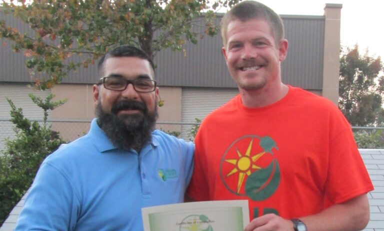 Two men smiling outdoors, one holding a certificate, with industrial building in background