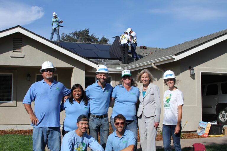 Team members in blue shirts stand in front of a house with solar panels being installed on the roof