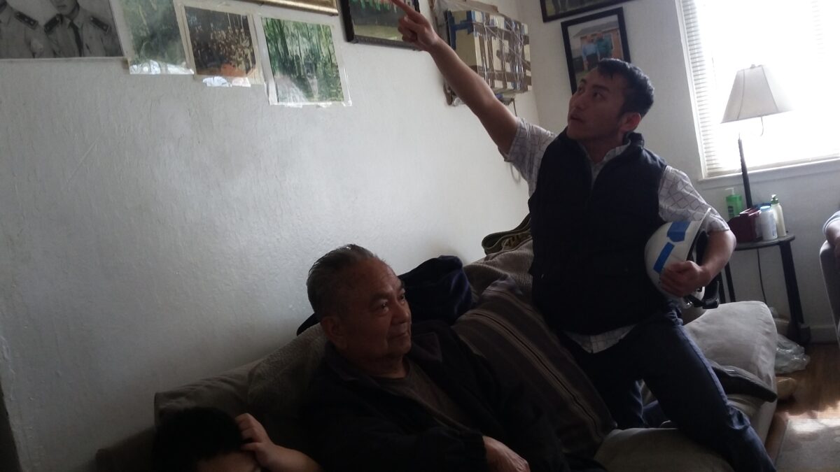 Two men sit on a couch in a living room; one points upward at framed photos on the wall while holding a ball
