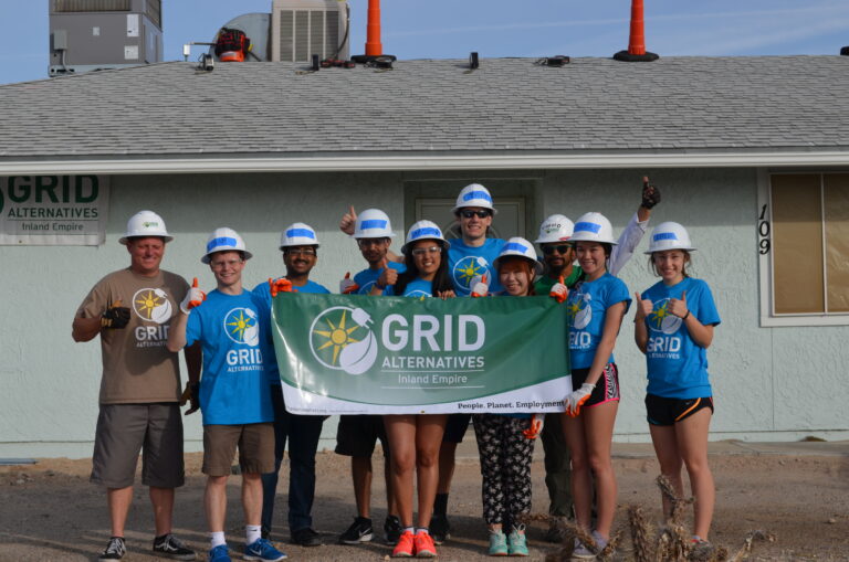 Duke students in hard hats hold GRID Alternatives Inland Empire banner in front of building