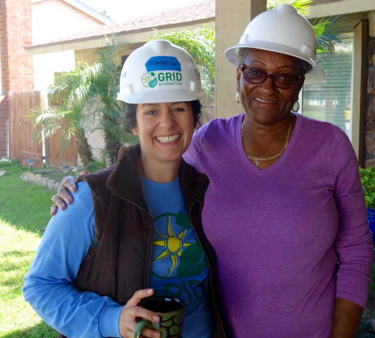 Two women smiling at camera wearing hard hats, one in Grid Alternatives shirt, standing in residential backyard