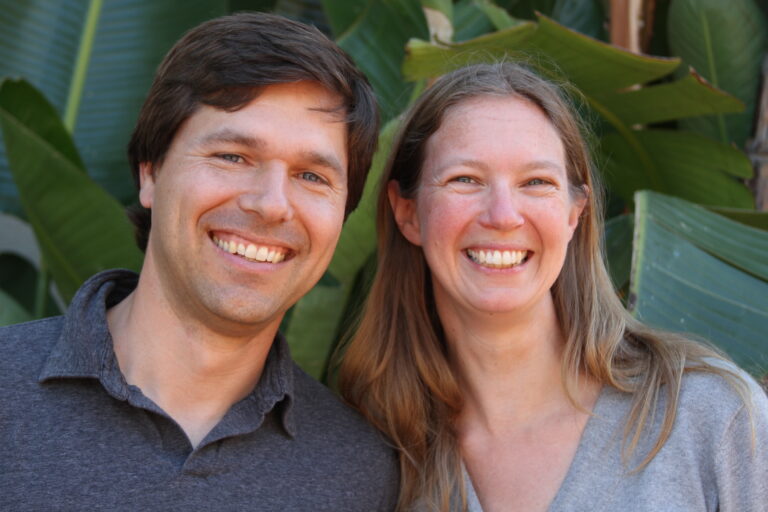 Two smiling people posing together in front of green foliage