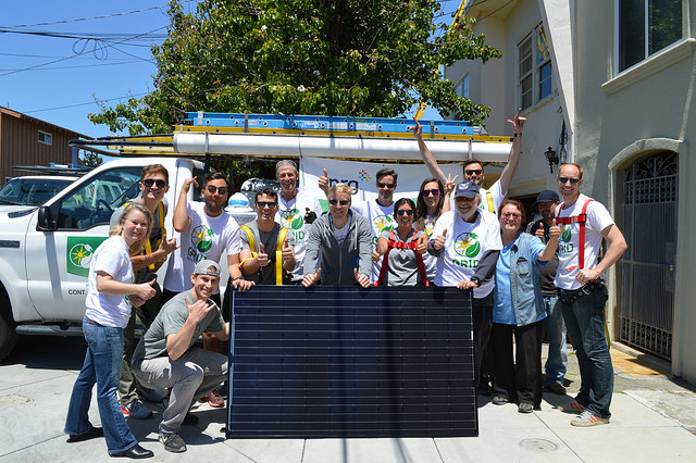 Group of people posing with a solar panel in front of a white van and residential building