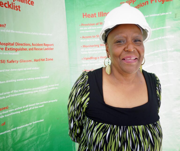 Woman in white hard hat smiling at camera in front of green "Heat Illness" safety presentation poster