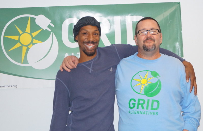 Two men smiling in front of a GRID Alternatives banner with sun and leaf logo
