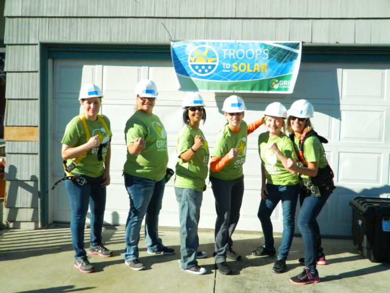 Six women in green shirts and hard hats pose in front of a "Troops to Solar" banner on a building