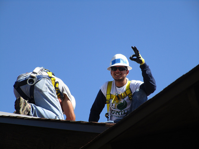 Construction worker on roof waving, wearing hard hat and safety gear against blue sky