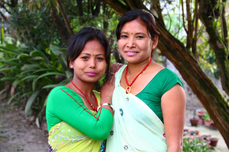 Two women in traditional Nepali attire smiling outdoors among green trees and foliage