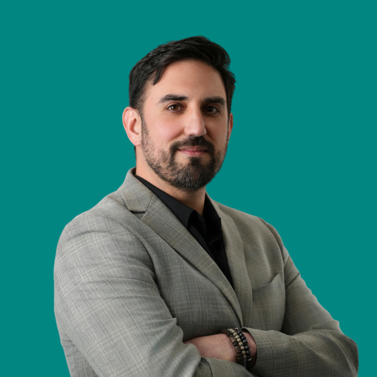 Professional headshot of a man with dark hair and beard wearing a gray blazer against a teal background