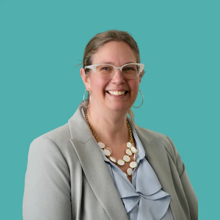 Erica Mackie, woman with glasses and braided hair wearing a gray blazer against a turquoise background