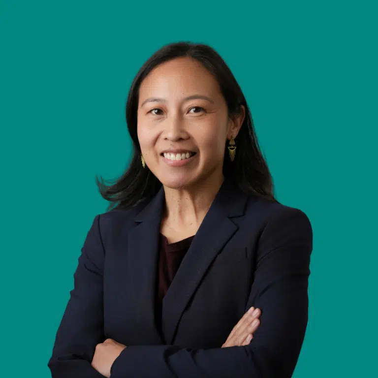 Professional headshot of Anna Bautista against a teal background, wearing a dark blazer with arms crossed, smiling at camera