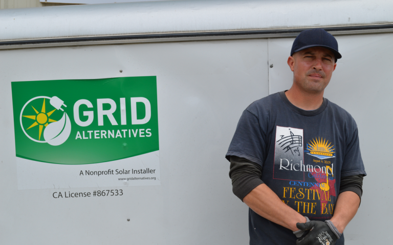 Solar installer wearing Grid Alternatives uniform stands in front of company van with green logo and California license number visible