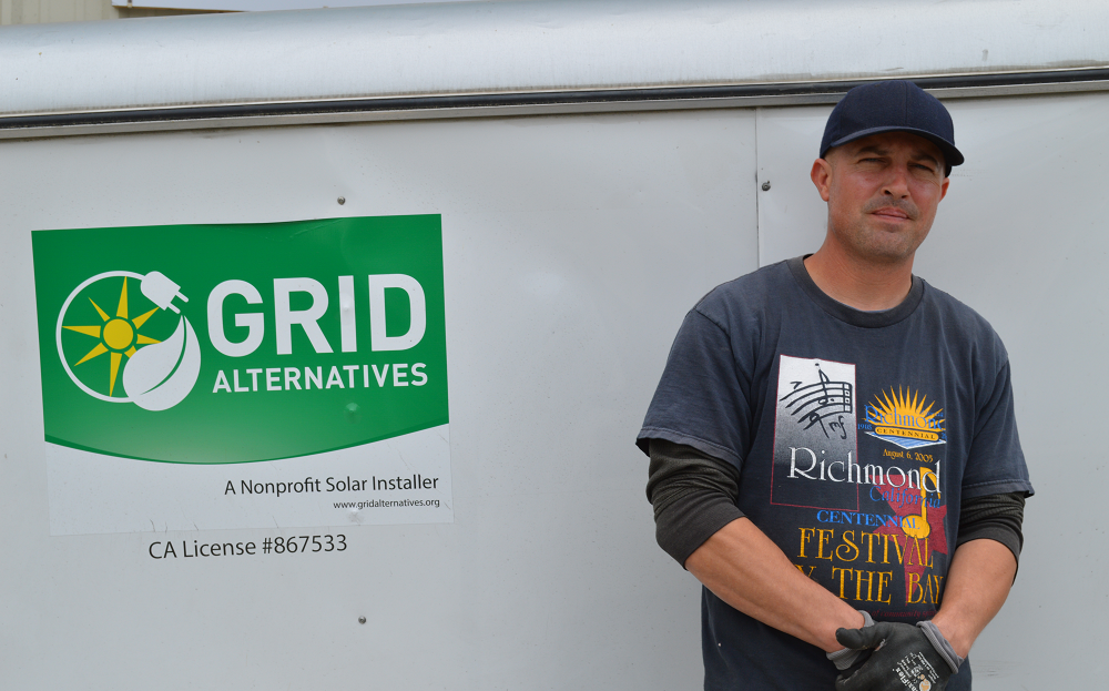 Solar installer wearing Grid Alternatives uniform stands in front of company van with green logo and California license number visible