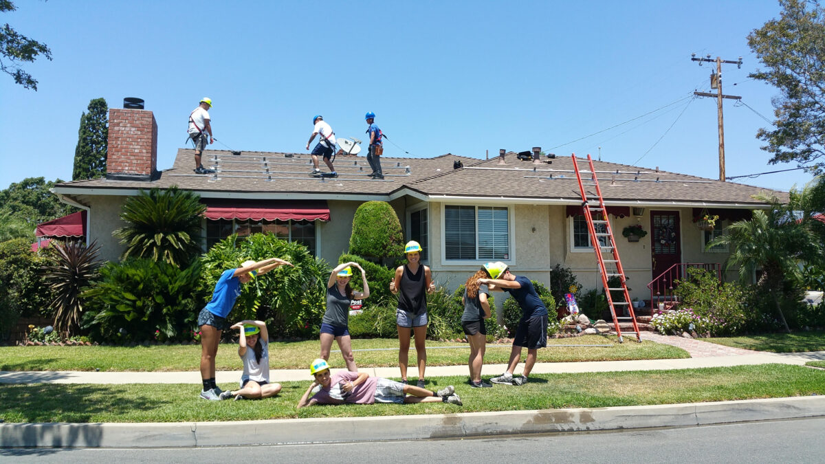 Community volunteers in hard hats working on roof repairs and exterior renovation of a residential house
