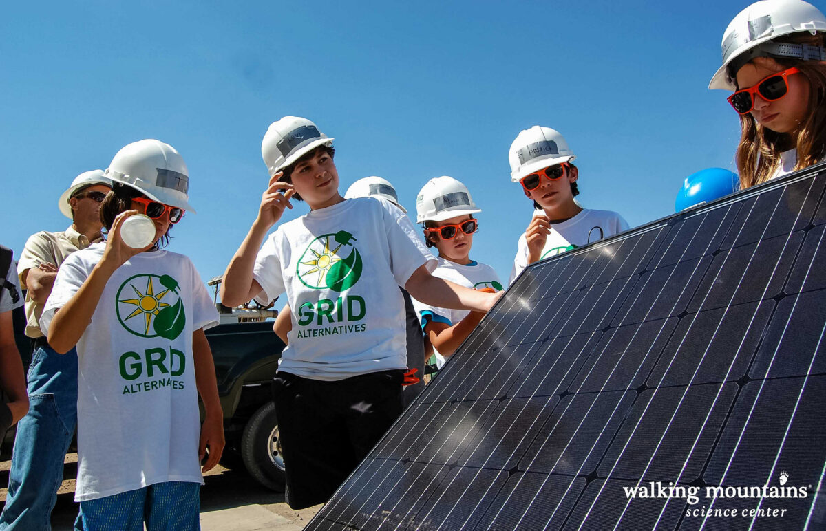 Group of students wearing hard hats and "GRID Alternatives" shirts examining solar panel installation