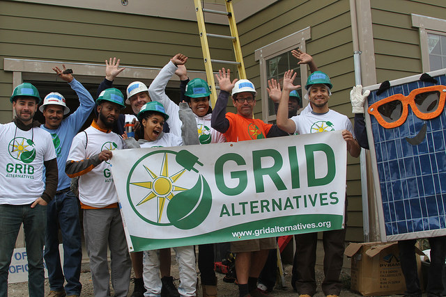 Team wearing hard hats holding GRID Alternatives banner with solar panel visible, celebrating renewable energy work
