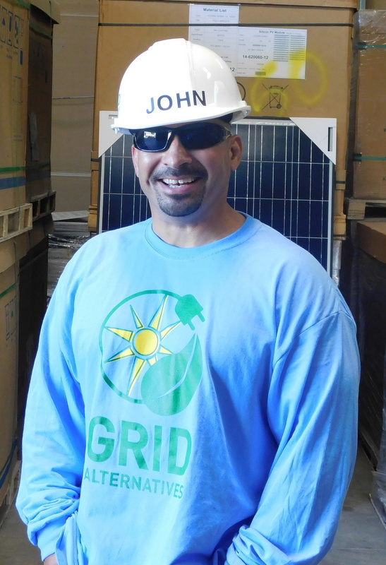 John wearing white hard hat and sunglasses, smiling in front of solar panel at GRID Alternatives warehouse