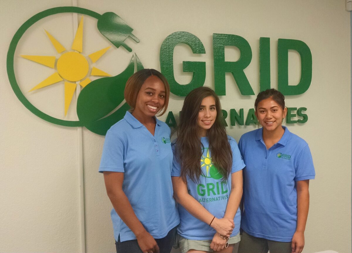 Three women in blue GRID Alternatives shirts stand in front of a GRID Alternatives logo featuring a sun and leaf design
