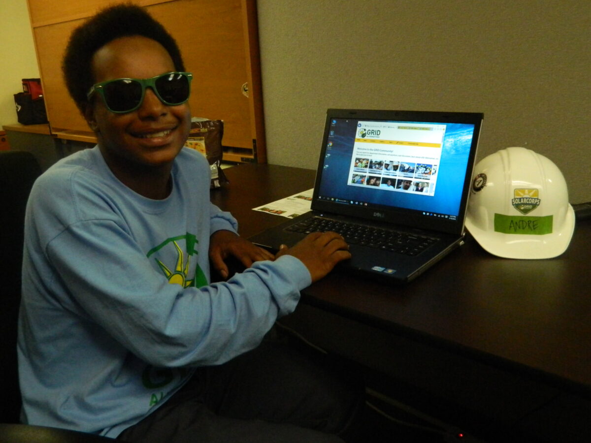 Person wearing sunglasses at desk with laptop displaying GRID website and white hard hat labeled "Andre