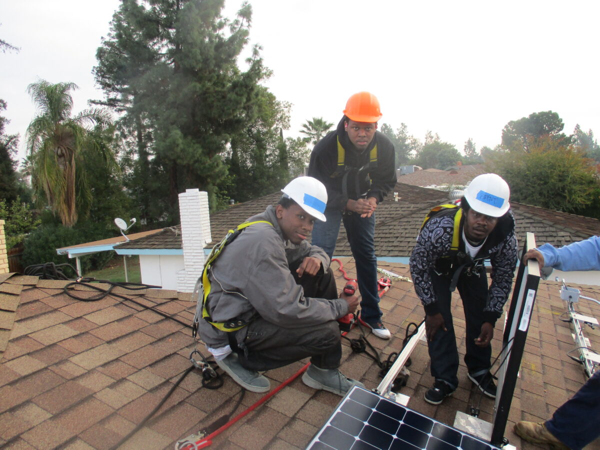 Four workers installing solar panels on a residential roof while wearing safety harnesses and hard hats